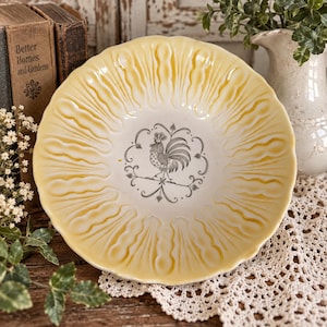 May include: A round ceramic bowl with a scalloped edge and a pale yellow glazed finish. The center of the bowl features a black line drawing of a rooster surrounded by a decorative circular vine pattern. The bowl has a textured, ribbed design.