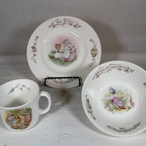 May include: A white ceramic set featuring Beatrix Potter illustrations, including a mug with a rabbit scene, a plate labeled Tom Kitten with a cat illustration, and a bowl labeled Jemima Puddle-Duck with a duck illustration, all with floral accents.