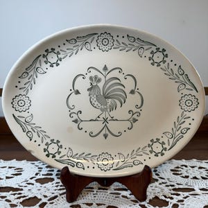 May include: An oval ceramic platter with a cream-colored base and a dark green printed design. The center features a rooster illustration surrounded by a decorative border of stylized flowers, leaves, and vines along the outer rim of the plate.