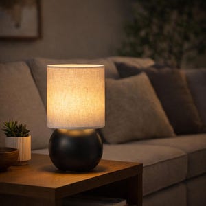 Könnte beinhalten: Eine kleine, moderne Tischlampe mit einem schwarzen, kugelförmigen Sockel und einem zylindrischen, beigefarbenen Stoffschirm. Die Lampe ist eingeschaltet und strahlt ein warmes Licht aus. Sie steht auf einem hölzernen Beistelltisch neben einer kleinen Topfpflanze, mit einem Sofa und Kissen im Hintergrund.