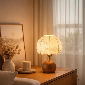 May include: A decorative table lamp with a floral patterned lampshade. The lamp has a wooden base and a brass stem. The lamp is on a wooden table next to a framed picture and a vase with dried flowers.