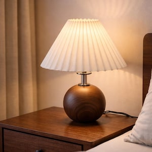 Puede incluir: Una lámpara de mesa de madera con base redonda y pantalla plisada blanca. La lámpara está sobre una mesita de noche marrón oscuro con un cajón. La lámpara está encendida, iluminando la habitación.