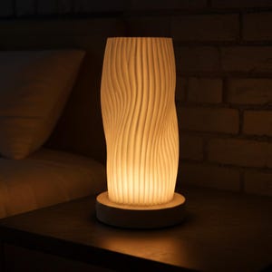 Puede incluir: Una lámpara de mesa texturizada de color crema con un diseño vertical y ondulado. La lámpara está iluminada, proyectando un brillo cálido. Se encuentra sobre una base redonda, colocada sobre una superficie oscura, con una pared de ladrillos al fondo.