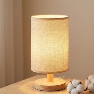 Pode incluir: Uma pequena luminária de mesa cilíndrica com uma tela de linho bege e uma base de madeira. A lâmpada está acesa, projetando um brilho quente. A base é redonda e feita de madeira clara. A lâmpada está em uma superfície de madeira.