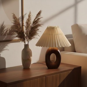 Puede incluir: Una lámpara de mesa de madera con una pantalla plisada de color crema y una base ovalada marrón oscuro. Un jarrón con hierba ornamental seca está al lado de la lámpara. La lámpara y el jarrón están sobre un mueble de madera.
