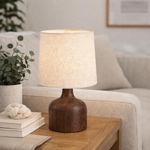May include: A table lamp with a dark wooden base and a beige linen shade. The lamp is on a wooden table, with a plant and books in the background. The lamp's design is simple and elegant, with a warm, neutral colour palette. The lamp is a stylish home decor item.