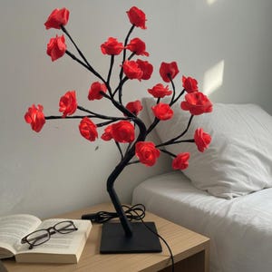 May include: A decorative lamp in the shape of a tree, featuring red rose blossoms. The black branches extend from a square base. The lamp is on a wooden bedside table next to an open book and spectacles.