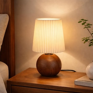 May include: A wooden table lamp with a round base and a pleated lampshade. The base is a sphere made of dark wood, and the shade is a light cream colour. The lamp is on a wooden bedside table.