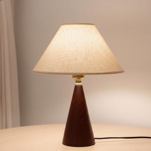 May include: A wooden table lamp with a conical base and a beige fabric lampshade. The base is dark brown, and the lampshade is a light beige colour. The lamp has a brass-coloured neck and a black power cord.