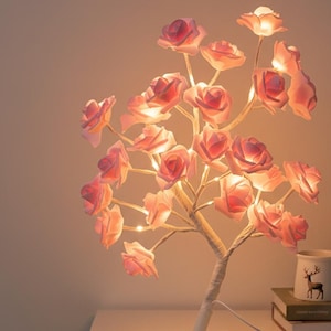 Puede incluir: Una lámpara decorativa de árbol de rosas con luces en forma de rosa rosa suave. El tronco del árbol está envuelto en tela blanca, y la lámpara está sobre una superficie blanca. Se ve una taza pequeña con una ilustración de ciervo.