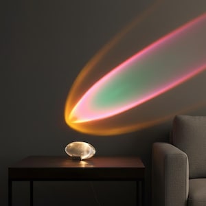 Peut inclure: Une lampe ovale moderne est posée sur une table en bois sombre, projetant un affichage lumineux coloré sur le mur. La lumière passe de l'orange au rose et au vert, créant une ambiance vibrante et artistique. Un canapé gris est partiellement visible.