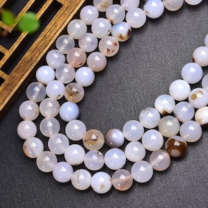 May include: Close-up of strands of white and light brown gemstone beads. The beads are spherical and have a polished finish. The beads are strung together, forming a necklace or bracelet. The background is a dark grey surface.