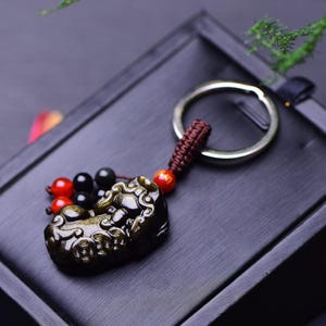 May include: A detailed, dark-coloured keychain pendant depicting a mythical beast. The pendant is a dark, golden-brown hue, with a braided brown cord and a silver keyring. Red and black beads are attached to the cord.