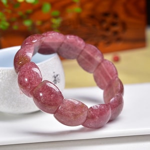 May include: A translucent, reddish-pink gemstone bracelet. The bracelet is composed of oval-shaped beads, each displaying a unique pattern of color variations. The beads are strung together, forming a circular shape. The bracelet is displayed on a white surface.