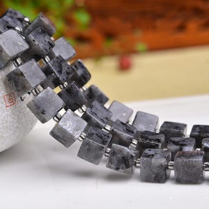 May include: A strand of grey labradorite cube beads with silver-coloured spacers. The beads are a mix of grey and black, with a slightly iridescent quality. The beads are strung together, creating a necklace or bracelet.