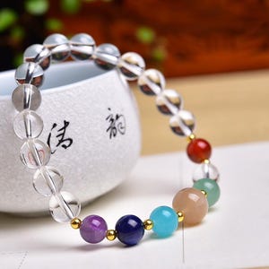 May include: A beaded bracelet featuring a variety of colorful gemstones and clear quartz beads. The bracelet includes purple, blue, turquoise, and peach-colored stones, along with a red bead and clear quartz spheres. The bracelet is displayed near a white ceramic cup.