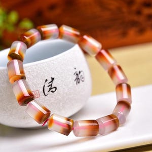 May include: A bracelet made of cylindrical beads in shades of brown, orange, and pink. The beads are strung together, forming a circular shape. The bracelet is resting on a white surface, with a white ceramic cup in the background.