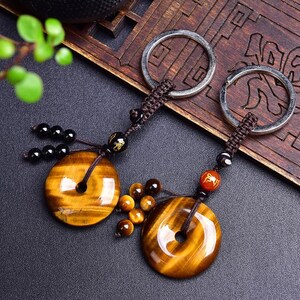 May include: Two keyrings featuring circular tiger's eye pendants. Each keyring has a silver ring, a brown woven cord, and decorative beads. The pendants are golden-brown with a polished finish. The background is dark with wooden accents.