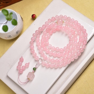 May include: A long strand of pink rose quartz beads, possibly a necklace or prayer beads, lies on a white surface. The beads are a consistent pink hue. A decorative tassel with a rose quartz pendant and green bead accents hangs from the strand.