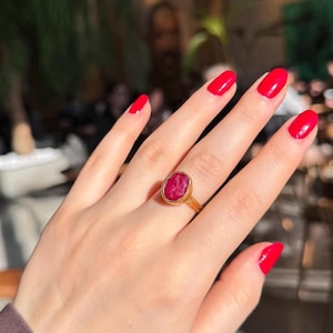 May include: A gold ring featuring a large, oval-cut, red gemstone. The ring is worn on a hand with red nail polish. The ring's setting is a thin, gold band. The gemstone has a slightly rough texture.