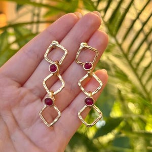 May include: A pair of gold-coloured drop earrings. Each earring features three connected geometric shapes: a square, a diamond, and another square. A small, round, red gemstone is set in the centre of each shape. The earrings are displayed against a blurred green background.