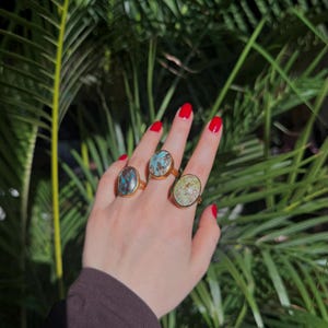 May include: Three gold rings, each with an oval gemstone in shades of blue, green, and brown. The rings are worn on a hand with red nail polish, with green foliage in the background.