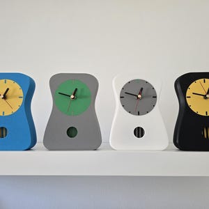 May include: Four contemporary clocks in blue, grey, white, and black. Each clock has an hourglass shape, a circular face with hands, and a pendulum-style cutout. The clock faces are yellow, green, grey, and yellow.