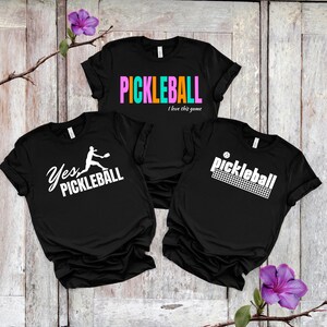 Può includere: Tre magliette nere con disegni a tema pickleball. Una maglietta presenta la parola "PICKLEBALL" in lettere colorate, un'altra dice "Yes, PICKLEBALL" con una silhouette di giocatore, e la terza mostra "pickleball" con una grafica a rete.