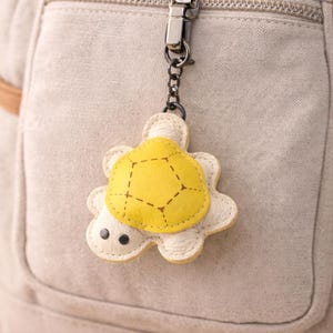 Puede incluir: Un llavero con forma de tortuga amarilla y blanca, colgado de un cierre metálico plateado. El llavero está sujeto a una bolsa beige con cremallera y un pequeño bolsillo. La tortuga tiene un caparazón amarillo y un cuerpo blanco.