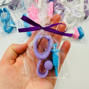 Puede incluir: Una bolsa de plástico transparente que contiene un jabón con forma de estetoscopio de color morado claro y un jabón con forma de jeringa de color azul claro, atada con una cinta morada oscura. Incluye una etiqueta blanca con el texto Thank You.