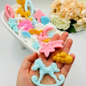 May include: A collection of colorful, decorative soap pieces in various shapes, including stars, crosses, flowers, and a rocking horse. The soaps are in shades of pink, blue, yellow, and white, arranged in a white dish and held in a hand.