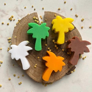 May include: Five palm tree-shaped soaps in white, green, yellow, orange, and brown colors. The soaps are arranged on a circular wooden slice with a visible grain pattern, surrounded by small scattered yellow particles on a light surface.