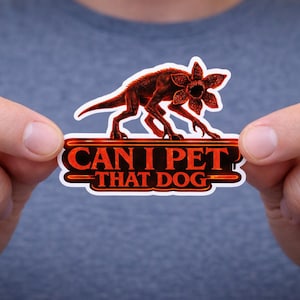 May include: A sticker featuring a Demogorgon-like creature with a flower-shaped head, in shades of red and brown. The text "CAN I PET THAT DOG" is displayed in red on a white and red background. The sticker is held between two hands.