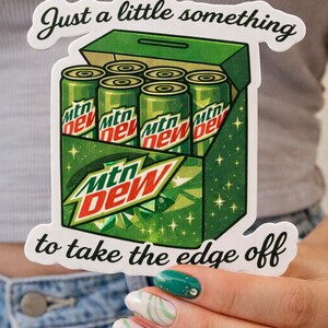 May include: A sticker depicting a green box containing six Mtn Dew cans. The box has the Mtn Dew logo and the text "Just a little something to take the edge off."