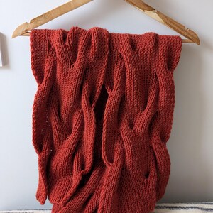 Könnte beinhalten: Eine rostfarbene Strickdecke mit Zopfmuster hängt an einem Holzkleiderbügel. Die Decke ist drapiert und zeigt die Textur und das Muster. Der Hintergrund ist eine neutrale Wand und ein gestreifter Stoff.