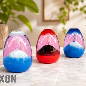 Peut inclure: Trois lampes à lave décoratives en forme d'œuf, bleues, rouges et bleues. Chaque lampe a une structure blanche en forme de montagne à la base, avec du liquide rose et bleu à l'intérieur. La base de chaque lampe est de couleur unie. Le mot "TEXON" est en bas.