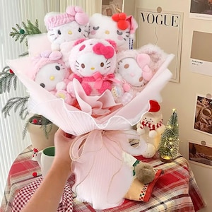 May include: A bouquet of plush Hello Kitty dolls in various pink outfits, wrapped in pink tissue paper and tied with a pink ribbon. The dolls have bows and headbands. The background includes a Vogue magazine cover and a small Christmas tree.