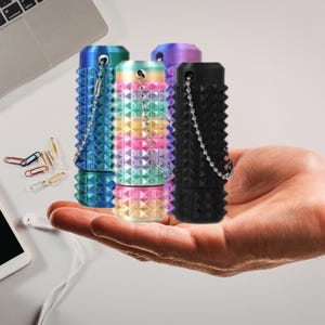 May include: Four colourful, cylindrical containers with a studded design are held in a hand. The containers are in shades of blue, rainbow, and black, each with a chain. Paper clips and a smartphone with earbuds are in the background.