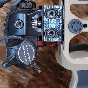 May include: Close-up of a black and tan bow sight with a black adjustment knob and a black and white label that reads "Mathews Genuine". The sight has a series of adjustment markings and is attached to a tan bow.