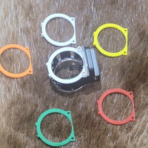 May include: An archery sight with a black and silver body, surrounded by five colored rings: white, yellow, orange, red, and green. The rings are circular with a square base. The sight is on a brown, textured surface.