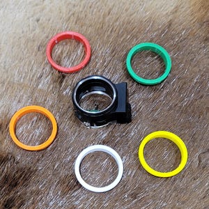 May include: A black archery sight with a clear lens is surrounded by six colored rings: red, orange, green, white, and yellow. The rings are arranged around the sight on a brown, textured surface.
