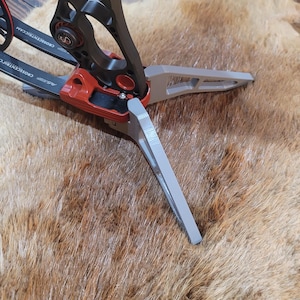 May include: A compound bow with a black frame and red accents. The bow features a gray stabilizer and is set against a brown fur background. The bow has the text "CROSSCENTRICCAM" printed on it.