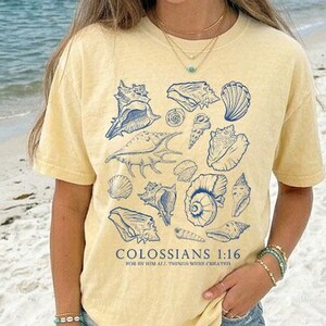 May include: A yellow t-shirt featuring a collection of blue seashell illustrations. The text "COLOSSIANS 1:16 FOR BY HIM ALL THINGS WERE CREATED" is printed below the seashells. The shirt is worn on a beach.