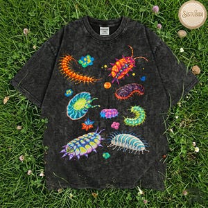 May include: A dark gray t-shirt featuring colorful illustrations of various insects and arthropods. The design includes a centipede, a pill bug, and other vibrant, detailed creatures. The shirt has a vintage, washed look.