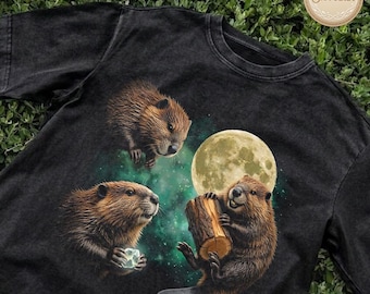May include: Black t-shirt featuring a whimsical illustration of three beavers. One beaver holds a log, another a glowing crystal, and the third is suspended above. A full moon and a starry green background complete the design.