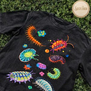 May include: Black t-shirt featuring colorful, whimsical illustrations of various insects and sea creatures. The design includes a centipede, pill bugs, and other unique, vibrant creatures. The shirt has a crew neck and short sleeves, perfect for casual wear.