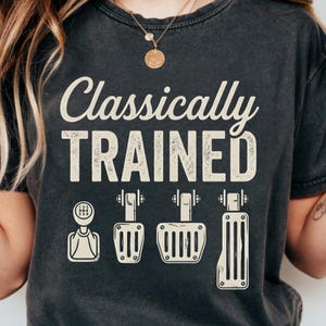 Może przedstawiać: Ciemnoszara koszulka z napisem "Classically TRAINED" w postarzanym, złamanym białym kolorze. Poniżej ilustracje dźwigni zmiany biegów i pedałów samochodowych. Koszulka ma wygląd vintage.