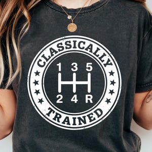 Może przedstawiać: Ciemnoszara koszulka z białym nadrukiem schematu zmiany biegów w okręgu. Tekst wokół okręgu brzmi "CLASSICALLY TRAINED" z gwiazdkami. Koszulka jest w stylu casual.