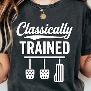 May include: Dark grey t-shirt featuring the white text "Classically TRAINED" above a graphic of three car pedals. The shirt has a vintage, worn appearance.