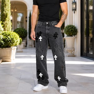 May include: A black crew neck t-shirt and charcoal gray denim jeans featuring white and black cross-shaped patches. The jeans have a straight-leg fit with decorative stitching patterns above the knee and a standard button and zipper fly closure.